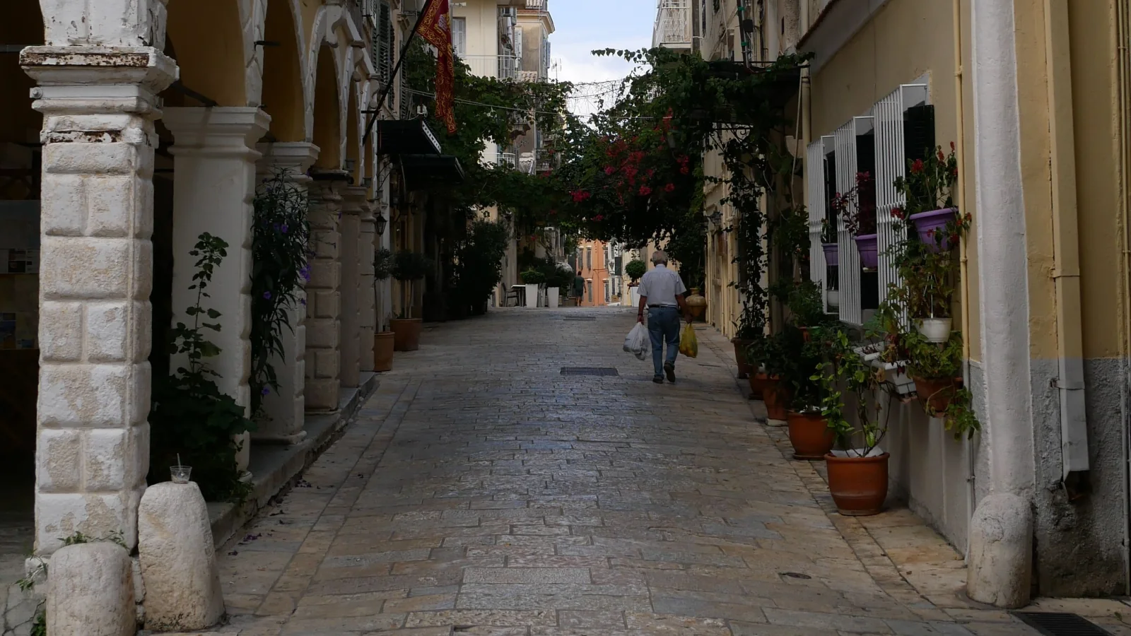 A pedestrian in a Corfu Town lane, with arcades and potted plants.