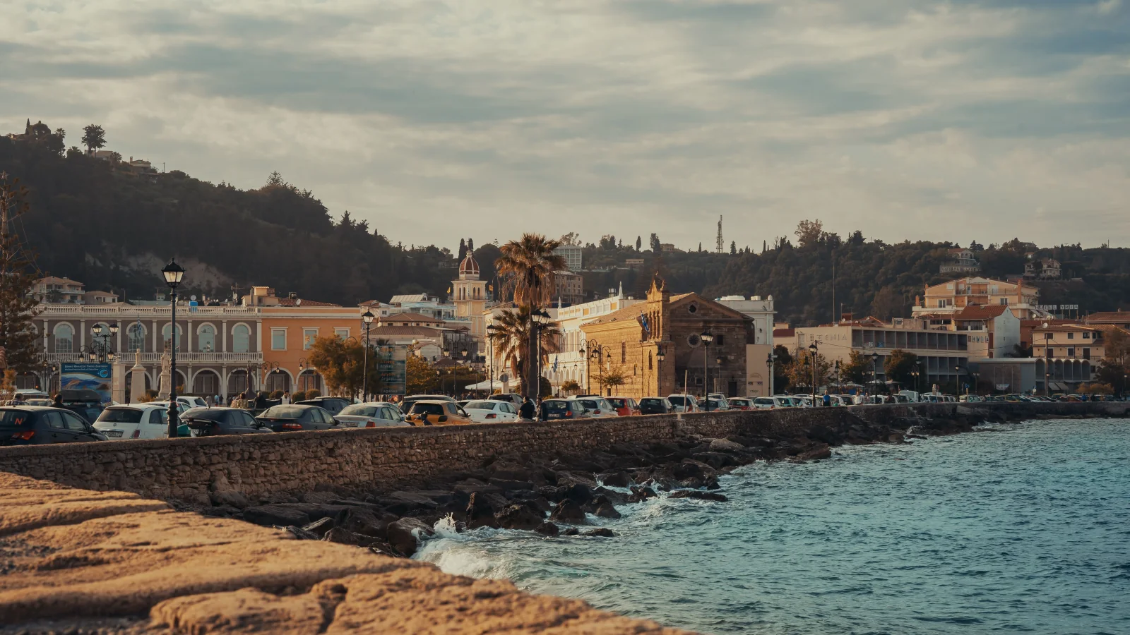 Zakynthos Town waterfront with buildings, parked cars and the wooded hillside behind.