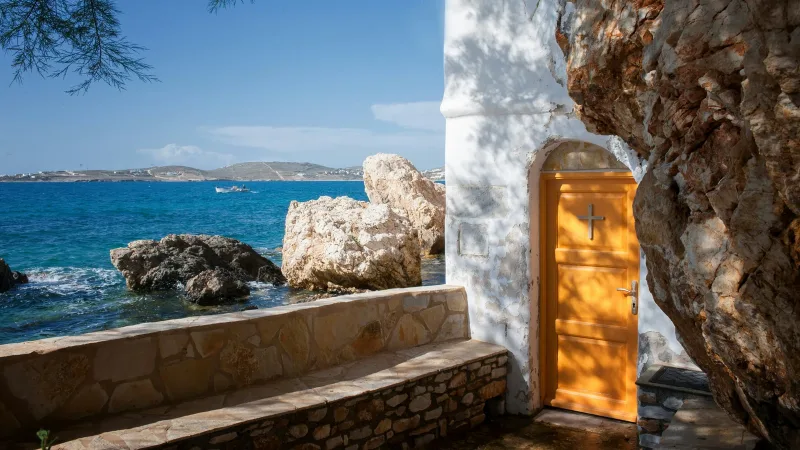 Small white coastal chapel with an orange door set into rock beside the Aegean Sea